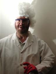 The professor wears a lab coat, goggles, and orange gloves.