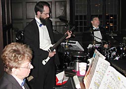 Ann plays piano in the foreground, James stands up to play behind the piano, and Jim is seated at his set in back.