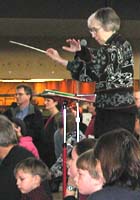 Carol conducts while some audience members sit on the edge of the stage.