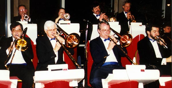 Four trombones are seated in front; four trumpets stand behind.