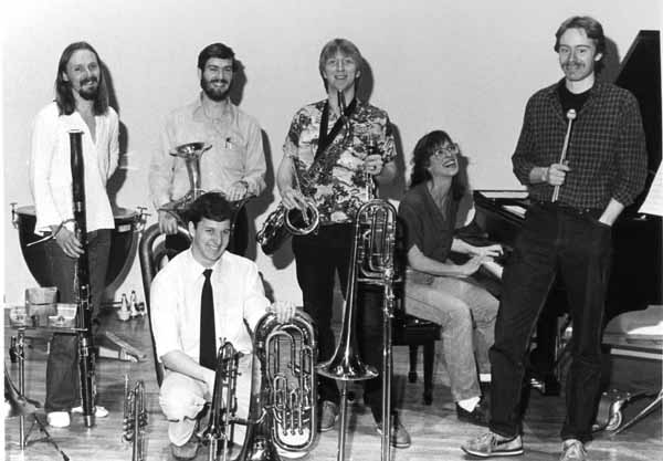 Jon kneels in front; Maggie sits at the piano; Dave E., Glen, Dave S., and Matt stand, surrounded by instruments used in the workshop.